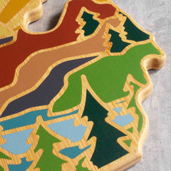 Introducing the Michigan Mitten Cutting Board with Artwork by Summer Stokes from Totally Bamboo: This decorative state-shaped cutting board showcases vibrant inlays of forests, rivers, and mountains against a gray backdrop. With its bamboo cutting surface, it seamlessly combines functionality with artistic elegance.