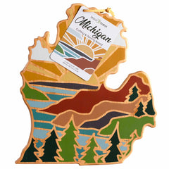 A Michigan Mitten Cutting Board designed by Summer Stokes, featuring a vibrant landscape design that includes mountains, trees, and the sun. The label at the top reads "Michigan Mitten Cutting Board" from Totally Bamboo and provides care instructions for maintaining its bamboo surface.