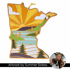 The Minnesota Cutting Board from Totally Bamboo features an illustration of the state shape adorned with a stylized landscape that includes a sun, trees, mountains, and a river in vibrant yellows, greens, and blues. Accompanying the design is text that reads "Artwork by Summer Stokes" with a circular inset photo next to it.