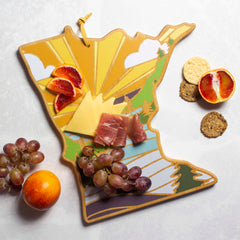 The Minnesota Cutting Board with Artwork by Summer Stokes from Totally Bamboo showcases charcuterie delights, including slices of cheese, cured meat, and blood orange, complemented by clusters of grapes. Nearby, you'll find crackers and a halved blood orange on a light surface. This distinctive piece seamlessly blends function with artistic flair to enhance your culinary presentation.