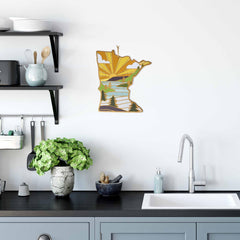 A modern kitchen with light gray cabinets, a black countertop, and a silver faucet showcases the Minnesota Cutting Board with Artwork by Summer Stokes from Totally Bamboo. A potted plant and a bowl rest on the counter, while a nature-themed wall decoration hangs above.