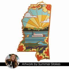 The Mississippi Cutting Board from Totally Bamboo features illustrated artwork by Summer Stokes, showcasing a sunburst over a riverboat surrounded by large flowers and a bluebird. The essence of Stokes' creativity shines through this design. A caption underneath reads: "Artwork by Summer Stokes," accompanied by her thumbnail and her dog.