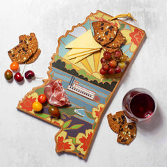 The Mississippi Cutting Board by Totally Bamboo, featuring artwork by Summer Stokes, serves as a vibrant backdrop for cheese, salami, grapes, and fruit slices beside a glass of red wine. The colorful board displays intricate patterns reminiscent of riverboats and floral designs. To enhance the experience, crackers are elegantly scattered around.