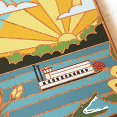 Illustrated scene depicting a steamboat on a blue river with the sun setting behind green hills. Stylized alligators and yellow flowers adorn the edges, capturing the Southern, rustic charm of Summer Stokes' artwork on the Mississippi Cutting Board by Totally Bamboo.