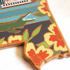 The Mississippi-shaped bamboo cutting board from Totally Bamboo features a delightful illustration by Summer Stokes, showcasing a vibrant green alligator with an open mouth, surrounded by large yellow and orange flowers on a colorful canvas.