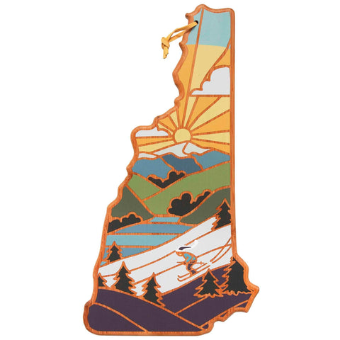 A depiction of a skier on a snowy mountain with stylized trees in the foreground and a sunrise over hills in the background, reminiscent of Summer Stokes' artwork, shaped like the state of New Hampshire. This design is ideal for Totally Bamboo's New Hampshire Cutting Board featuring artwork by Summer Stokes.