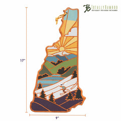 The Totally Bamboo New Hampshire Cutting Board, designed by Summer Stokes, showcases an elegant engraving of a sunlit landscape with mountains and a skier. Crafted from bamboo, it measures 17 by 9 inches and features the logos of both Totally Bamboo and artist Summer Stokes.