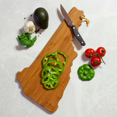 Sliced green bell peppers rest on the New Hampshire Cutting Board with Artwork by Summer Stokes, by Totally Bamboo. Beside it lie a knife, a whole green pepper, tomatoes, an avocado, garlic, basil, and peppercorns on a light surface.