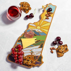 The New Hampshire Cutting Board with Artwork by Summer Stokes from Totally Bamboo features a distinctive shape that resembles the state's outline, elegantly displaying slices of cheese, cured meat, almonds, and seeded crackers. Fresh cherries and a cluster of grapes are also thoughtfully arranged nearby. A glass of red wine accompanies the setup on a light surface.