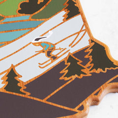 Illustration of a skier gracefully descending a snowy mountain slope surrounded by pine trees, beautifully bordered by a wooden frame resembling the New Hampshire Cutting Board with Artwork by Summer Stokes from Totally Bamboo, capturing the spirit of the state.