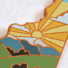 The New Hampshire Cutting Board by Totally Bamboo features a Summer Stokes artwork depicting a sun rising over mountains, with stylized rays and clouds accented in blue, green, yellow, and brown hues. This wooden piece boasts a natural, textured look against a light background, merging the practicality of a cutting board with artistic elegance.