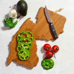 The New Jersey Cutting Board with Artwork by Summer Stokes from Totally Bamboo beautifully captures the Garden State's silhouette while displaying sliced green bell peppers. Nearby, a knife lies amongst vine-ripened tomatoes, a whole pepper, an avocado, garlic cloves, and fresh basil leaves on a pristine white surface.