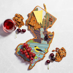 The New Jersey Cutting Board with Artwork by Summer Stokes, from Totally Bamboo, features a vibrant design of nature in its colorful artwork. It beautifully holds an assortment of cheese, almonds, cherries, and grapes. Accompanied by seeded crackers and a glass of red wine on the marble surface nearby, this bamboo cutting board is both functional and artistic.