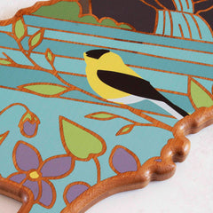 The New Jersey Cutting Board by Totally Bamboo showcases artwork by Summer Stokes, featuring a vibrant design of a yellow bird perched on a branch adorned with leaves and purple flowers against a blue backdrop. The edge of the artwork has a scalloped pattern.