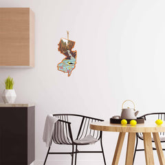 The modern kitchen features a wooden cabinet, a black wire chair, and a round table decorated with lemons and a teapot. A vibrant state map artwork by Summer Stokes embellishes the white wall. On the counter, there's a geometric plant pot next to a New Jersey Cutting Board with artwork by Summer Stokes from Totally Bamboo.