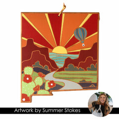 The New Mexico Cutting Board by Totally Bamboo features vibrant artwork by Summer Stokes, highlighting a southwestern sunset with a hot air balloon, river, cacti, and mountains. A person and a dog are depicted in a circular inset. The text on the board reads "Artwork by Summer Stokes," reflecting her distinctive style often seen on state-shaped cutting boards.