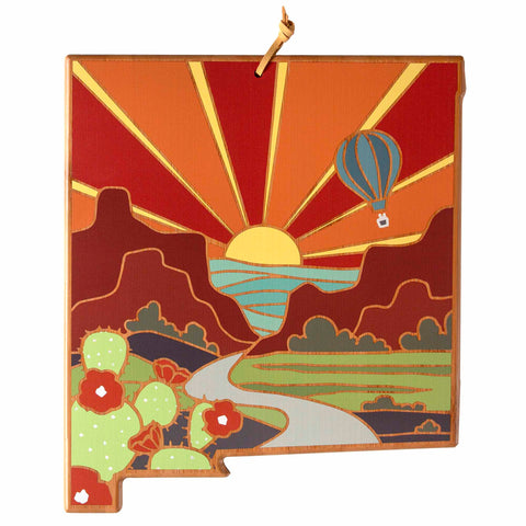 The New Mexico Cutting Board by Totally Bamboo features a vibrant artwork by Summer Stokes, depicting a sunrise over desert mountains, a winding river, and a hot air balloon in the sky. In the foreground, there are cacti adorned with red flowers. The artwork showcases colorful shades of red, orange, green, and yellow across the landscape.