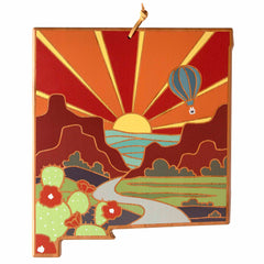 The New Mexico Cutting Board by Totally Bamboo features a vibrant artwork by Summer Stokes, depicting a sunrise over desert mountains, a winding river, and a hot air balloon in the sky. In the foreground, there are cacti adorned with red flowers. The artwork showcases colorful shades of red, orange, green, and yellow across the landscape.