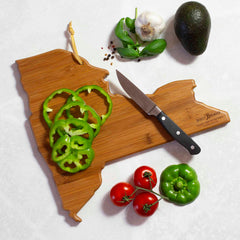 A New York-shaped cutting board, crafted from bamboo by Totally Bamboo and featuring artwork by Summer Stokes, showcases sliced green bell pepper, a paring knife, tomatoes, a whole bell pepper, avocado, garlic, basil, and red pepper flakes on a pristine white surface.