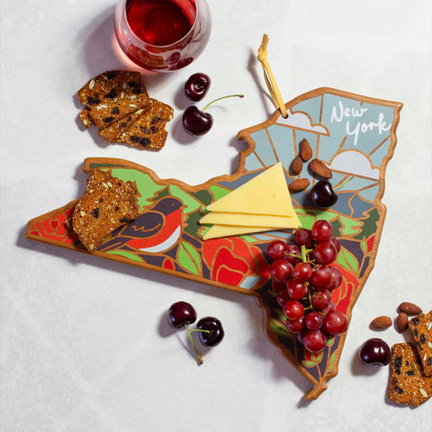 The New York Cutting Board with Artwork by Summer Stokes from Totally Bamboo elegantly displays cheese slices, grapes, cherries, and nuts. Several crackers are scattered around the board. Next to it, a glass of red liquid rests on the white surface.