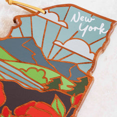 Crafted in the shape of New York, this decorative wooden cutting board from Totally Bamboo features a vibrant summer landscape with mountains and sun rays. The design includes "New York" elegantly inscribed at the top in cursive, reflecting the artistic style of Summer Stokes Artwork.