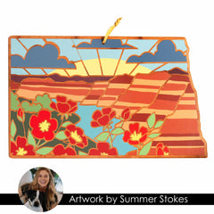 The North Dakota Cutting Board with Artwork by Summer Stokes from Totally Bamboo showcases a vibrant landscape featuring red flowers, colorful hills, and a sunrise with rays and clouds. This rectangular piece includes a hanging loop for display, reminiscent of State-Shaped Cutting Boards.