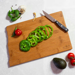 On a North Dakota Cutting Board with artwork by Summer Stokes, a knife is surrounded by fresh ingredients including sliced green bell pepper, whole bell pepper, cherry tomatoes, garlic, basil, black peppercorns, and an avocado on a marble background. This scene encapsulates the vibrant essence of Summer Stokes' artistry with fresh ingredients poised for creative culinary endeavors. Brought to you by Totally Bamboo.