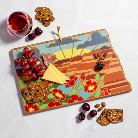 The North Dakota Cutting Board by Totally Bamboo, adorned with Summer Stokes' floral and landscape designs, displays a spread of grapes, cheese, chocolate, nuts, and crackers. A striking bamboo surface is complemented by a nearby glass of red liquid and scattered cherries.