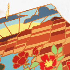 The North Dakota Cutting Board by Totally Bamboo, featuring artwork by Summer Stokes, beautifully captures a sunset over a striped canyon landscape adorned with clouds above. Vibrant red and yellow flowers grace the foreground, while an elegant gold ribbon in the top left corner adds a sophisticated touch to this colorful masterpiece.