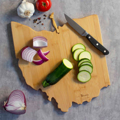 An Ohio Cutting Board with Artwork by Summer Stokes, crafted by Totally Bamboo, holds sliced zucchini, red onion pieces, and a paring knife. Nearby on the gray surface are a whole zucchini, garlic, a tomato, and peppercorns—a harmonious blend of utility and artistry reminiscent of Summer Stokes' creations.