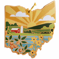 The Ohio Cutting Board with Artwork by Summer Stokes, produced by Totally Bamboo, features a captivating rural scene. The bamboo surface depicts an illustration of the state of Ohio, complete with a barn amid rolling fields and vibrant yellow flowers beneath a sunset sky pierced by sun rays.