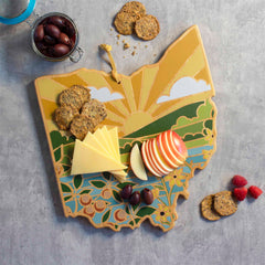 The Ohio Cutting Board with Artwork by Summer Stokes from Totally Bamboo is adorned with a landscape design and displays sliced apples, cheese, crackers, and black olives. Additional crackers are scattered on the gray surface alongside a small cluster of raspberries.