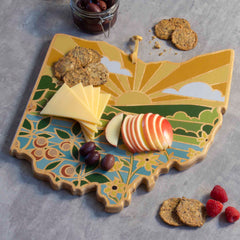 An eye-catching Ohio-shaped bamboo cutting board from Totally Bamboo, featuring Summer Stokes' bright depiction of a sunny landscape, elegantly displays cheese slices, apple slices, olives, and crackers. A small jar of olives and some scattered crackers complement the stylish gray backdrop.