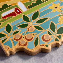 Close-up of a vibrant Ohio cutting board from Totally Bamboo, featuring artwork by Summer Stokes that depicts a rural scene with a barn, sunflowers, and rolling hills. The wavy edge of the board adds an artistic touch as it rests on a gray surface.