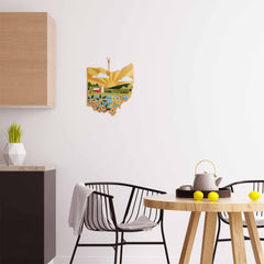 A modern kitchen features a black chair and a round wooden table adorned with a teapot and lemons. A state-shaped Ohio Cutting Board with Artwork by Summer Stokes from Totally Bamboo complements the minimalist decor, which includes a potted plant and geometric objects on the bamboo cutting surface.