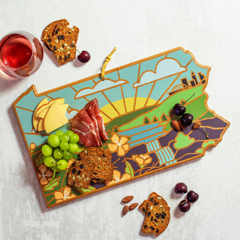 The Pennsylvania Cutting Board with Artwork by Summer Stokes, from Totally Bamboo, is elegantly adorned with meats, cheese, grapes, and crackers. Nearby, a glass of rosé sits on the white bamboo surface alongside cherries and extra crackers.