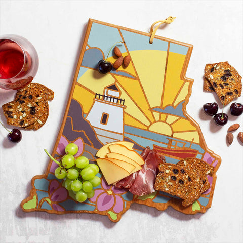 The Rhode Island Cutting Board by Totally Bamboo, showcasing the vibrant lighthouse and sunset design by Summer Stokes, is elegantly adorned with grapes, cheese slices, cured meats, seeded crackers, almonds, and cherries as a delightful snack arrangement. Accompanying this spread is a stemless glass of red wine.