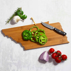 The Tennessee Cutting Board with Artwork by Summer Stokes from Totally Bamboo features sliced green bell peppers and a knife, while nearby, a bunch of tomatoes, halved red onion, garlic, basil, and a sprig of rosemary are arranged on a light surface.