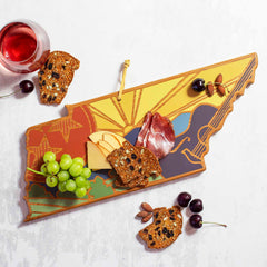 A Tennessee Cutting Board with Artwork by Summer Stokes, crafted from Totally Bamboo, beautifully displays grapes, cheese slices, cured meat, and crackers, along with scattered cherries and almonds. Nearby, a glass of red wine enhances the vibrant design featuring a guitar and sunburst motif—pure Summer Stokes artwork.