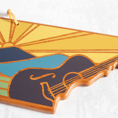 A Tennessee cutting board by Totally Bamboo features a vibrant Summer Stokes artwork, showcasing a sunrise over mountains. The image combines shades of yellow, blue, and green with a glossy finish and offers the functionality of a hanging loop at the top.