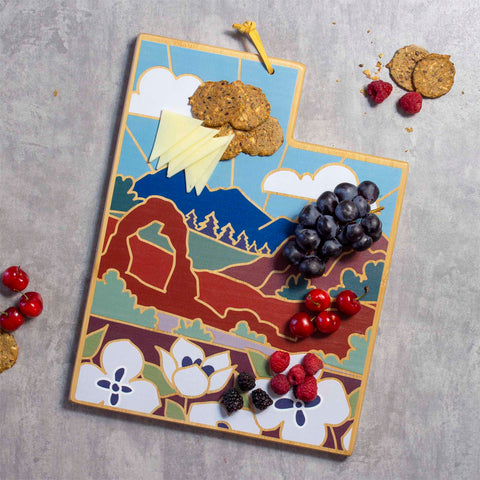 The Utah Cutting Board with Artwork by Summer Stokes, crafted from bamboo by Totally Bamboo, showcases vibrant landscape designs of mountains and arches. It's elegantly arranged with cheese slices, crackers, grapes, raspberries, blackberries, and cherries on a textured gray surface.