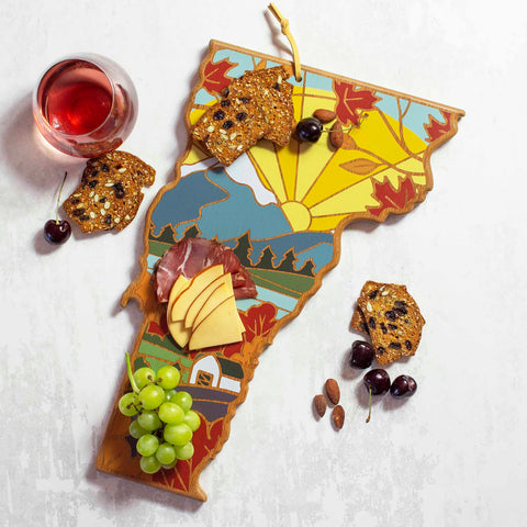 The Vermont Cutting Board with Artwork by Summer Stokes from Totally Bamboo features a vibrant design, making it ideal for arranging slices of cheese, prosciutto, grapes, and more. Beside it rests a glass of rosé wine on the white surface surrounded by scattered cherries and almonds.