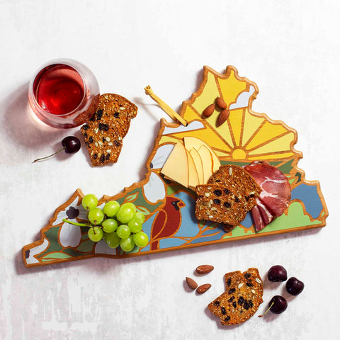 The Virginia Cutting Board with Artwork by Summer Stokes, a product by Totally Bamboo, features a vibrant state map design. It beautifully presents an assortment of cheese, cured meats, crackers, almonds, grapes, and cherries. A glass of pink wine sits nearby on the light surface, enhancing the charm of the scene.