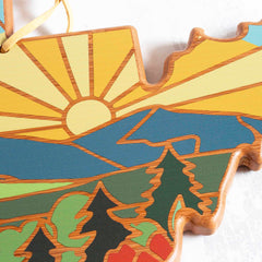 A wooden cutting board by Totally Bamboo, shaped like the state of West Virginia, showcases Summer Stokes' artwork featuring a stylized landscape with a sun rising over mountains, pine trees, and a colorful sky.