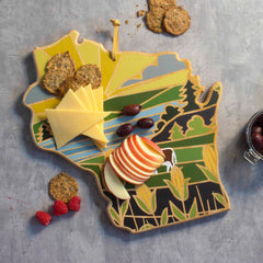 The Wisconsin Cutting Board with Artwork by Summer Stokes, from Totally Bamboo, is a bamboo cutting surface shaped like Wisconsin, featuring cheese, apple slices, olives, and crackers enhanced by the vivid artistry of Summer Stokes. Crackers and raspberries are elegantly arranged on the gray background.