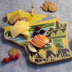 A Wisconsin-themed centerpiece, the Totally Bamboo Wisconsin Cutting Board with Artwork by Summer Stokes, is adorned with sliced cheese, apple slices, grapes, and crackers. It features illustrations of fields and a sky and is complemented by raspberries scattered nearby.