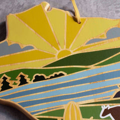 Introducing the Wisconsin Cutting Board with Artwork by Summer Stokes from Totally Bamboo. This wooden piece beautifully captures a sunrise over green hills and a blue river, adorned with stylized trees in the distinctive shape of Wisconsin. It comes equipped with a convenient string for hanging, reminiscent of Summer Stokes' renowned style.