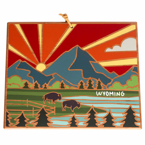 The Wyoming Cutting Board with Artwork by Summer Stokes, crafted by Totally Bamboo, showcases a vibrant illustration of a Wyoming landscape. It beautifully depicts a mountain at sunset, two bison grazing beside the lake, and trees in the foreground. The sky is adorned with a cloud and rays of sunlight, while "Wyoming" is elegantly scripted near the bison.