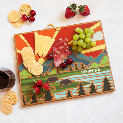 A lively Wyoming Cutting Board with Artwork by Summer Stokes from Totally Bamboo features a picturesque Wyoming landscape with mountains, trees, and bison. Topped with cheese, salami, grapes, cherries, strawberries, and crackers, it beckons a nearby glass of red wine. Enjoy this stunning tribute to state-shaped cutting boards.