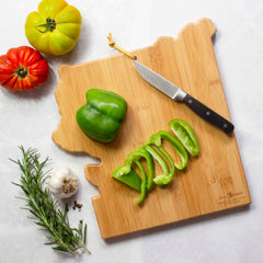 The Yellowstone Cutting Board with Artwork by Summer Stokes from Totally Bamboo features a beautifully designed bamboo cutting surface, showcasing sliced and whole green bell peppers alongside a knife. Nearby, you'll find two tomatoes, a garlic bulb, a rosemary sprig, and peppercorns.